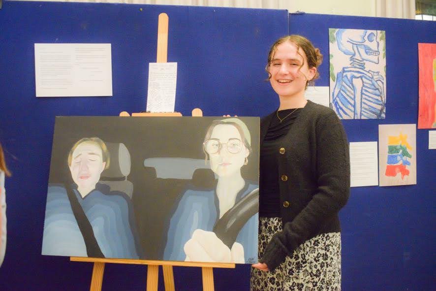 Yael Aliza smiling beside her painting I’ll drive on an easel at an exhibition